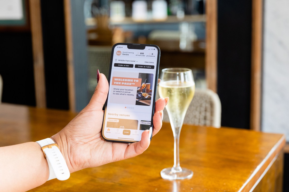 Woman holding up a smartphone with The Pass app open, with a glass of champagne in the background on a table.