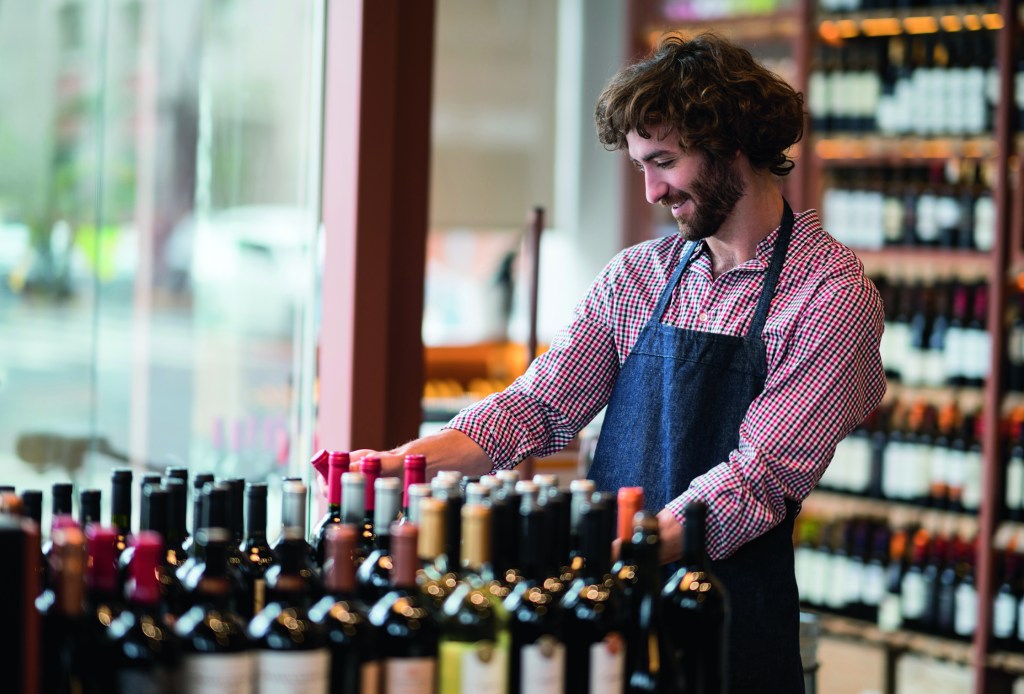 Young employee at a wine store organizing the bottles on a stand looking happy and smiling