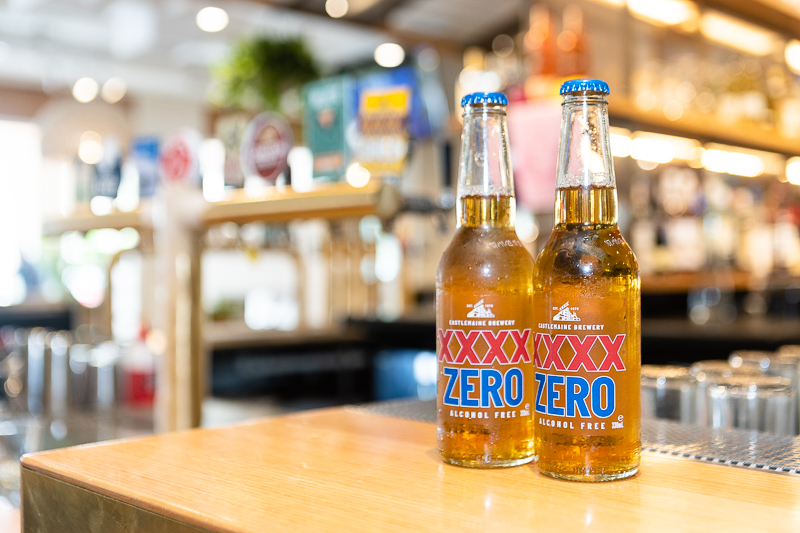 two bottles of XXX Zero stand upright in the foreground on a wooden bar