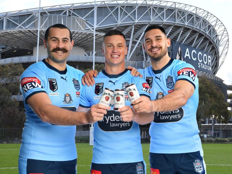 Three rugby players dressed in the NSW Rugby League jerseys stand outside a stadium, holding up cans of Jim Beam RTDs.