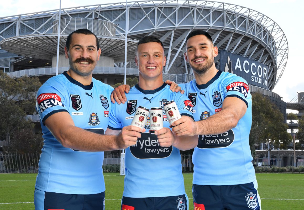 Three rugby players dressed in the NSW Rugby League jerseys stand outside a stadium, holding up cans of Jim Beam RTDs.