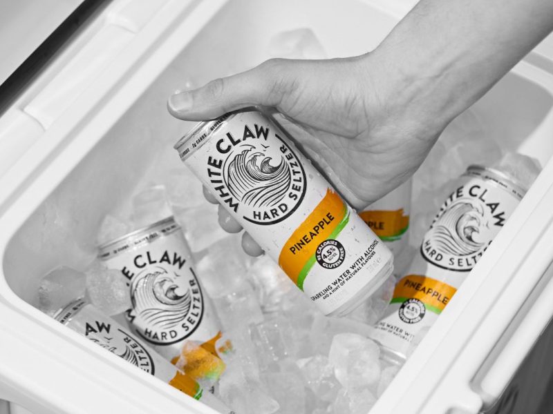 A hand reaches into an icebox to grab a can of White Claw Pineapple, which is white with orange accents.