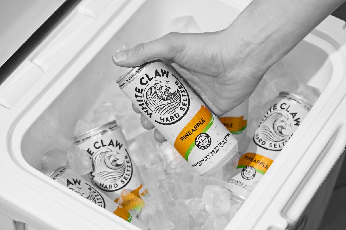 A hand reaches into an icebox to grab a can of White Claw Pineapple, which is white with orange accents.